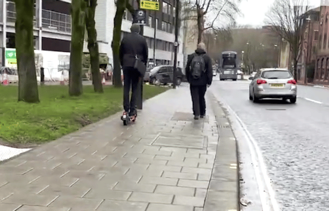 An e-scooter rider is caught in the video riding on the pavement, which is not allowed, precariously close to a pedestrian.
